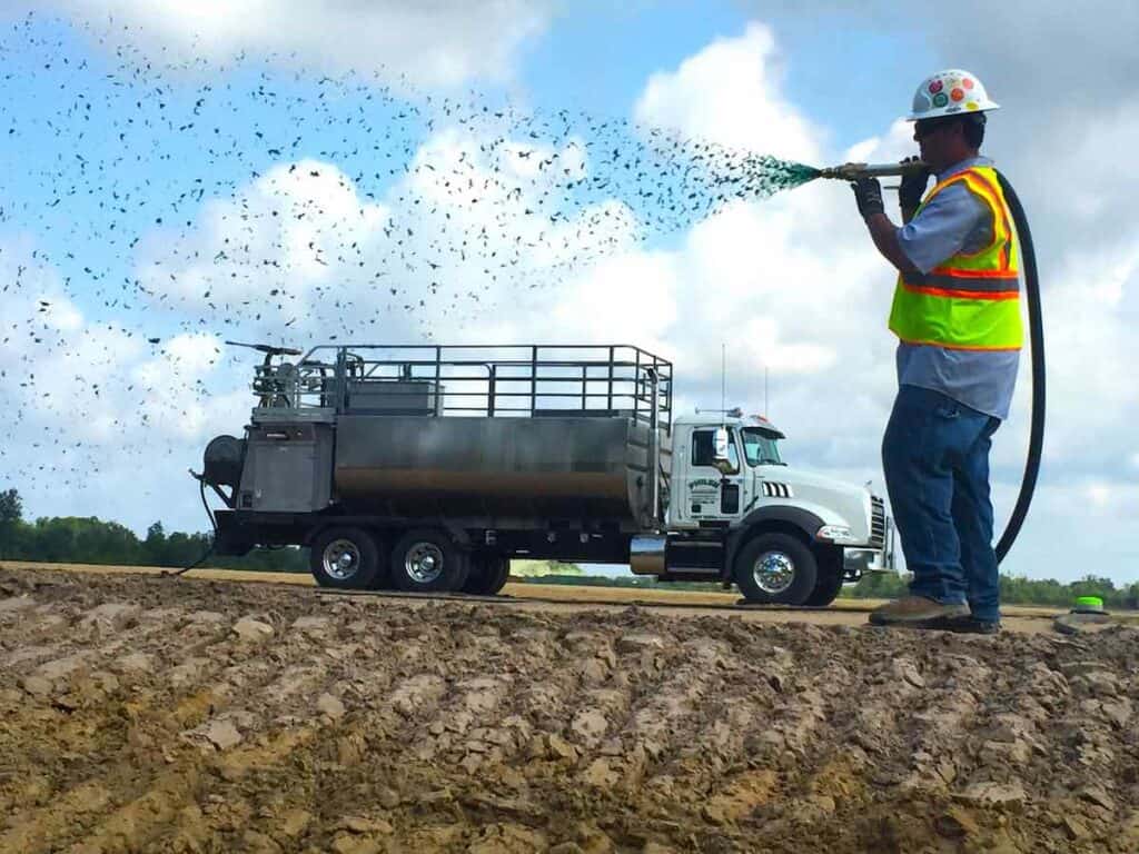 commercial hydroseeding costs. A person holding a hydroseeding nozzle over their shoulder sprays hydroseed over bare dirt. A hydroseeder is visible parked in the background.
