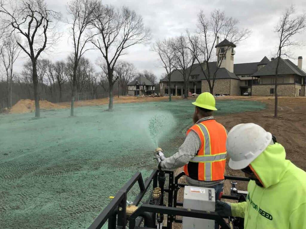 Environmental Benefits of Hydroseeding. A crew of two people stand on top of a hydroseeder, one controls a nozzle spraying green hydroseed over a bare field.