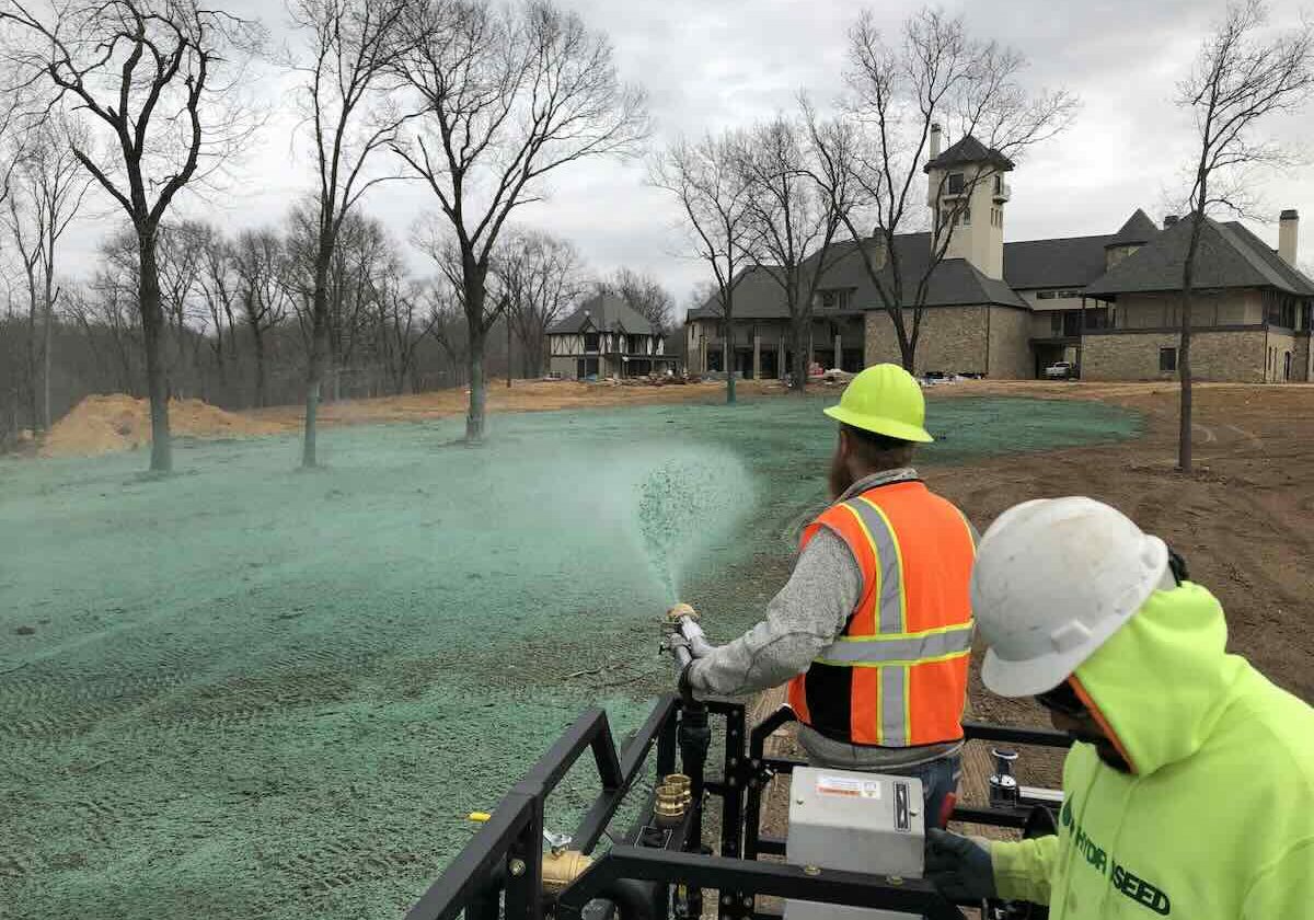 Environmental Benefits of Hydroseeding. A crew of two people stand on top of a hydroseeder, one controls a nozzle spraying green hydroseed over a bare field.